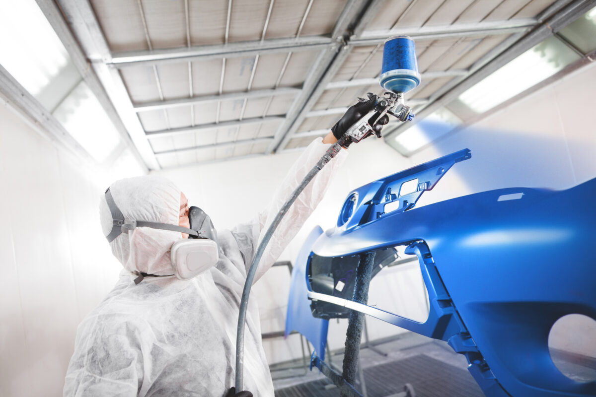 Male worker in protective clothes and mask painting car bumper using spray paint.